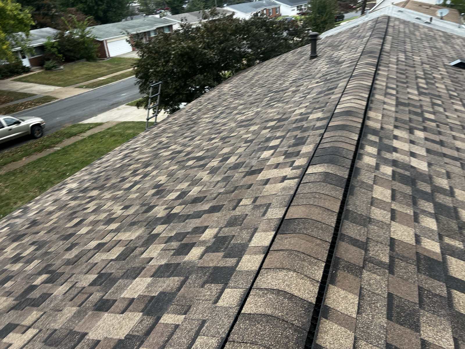 The capped ridge of a newly laid brown, tan and charcoal architectural shingle roof running diagonally across the frame with a dark ridge vent beneath the caps, an aluminium ladder hooked over the eave at the left, and a street of single storey brick and vinyl ranch houses with a white pickup parked below